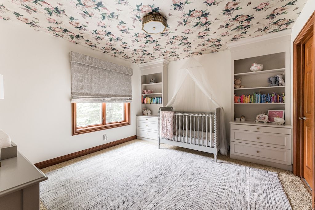 Nursery room with floral ceiling wallpaper, crib, built-in shelving, window, and light-colored rug.
