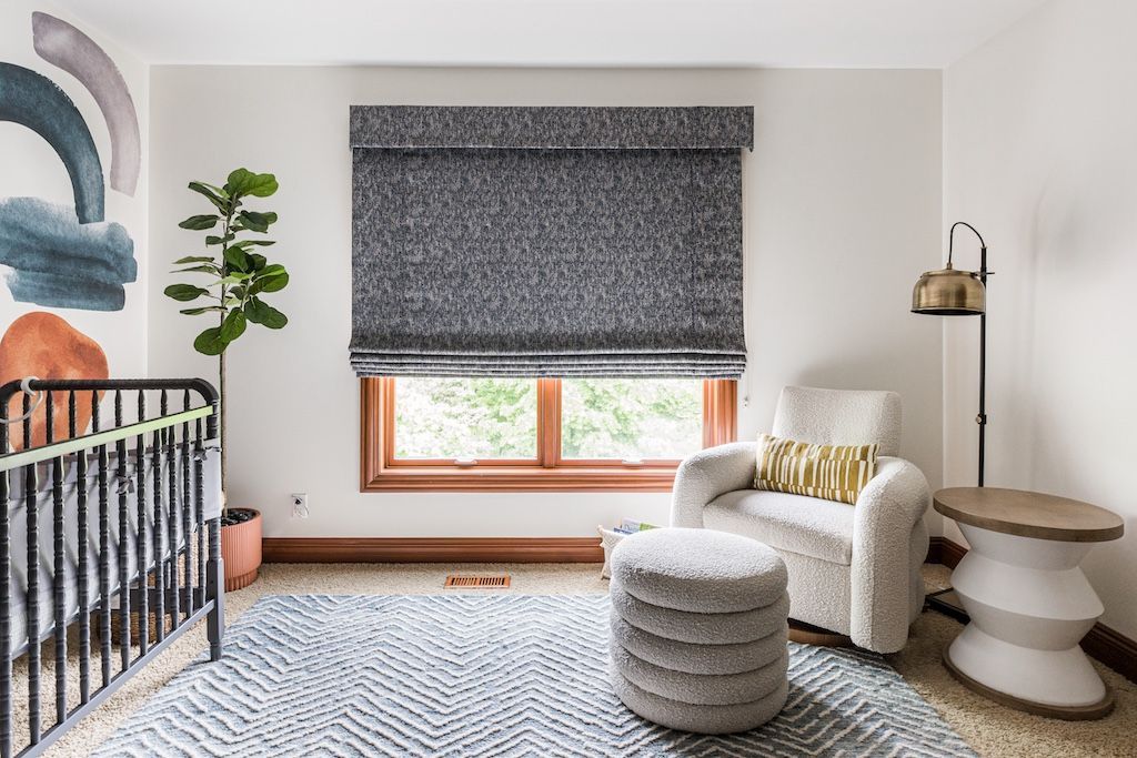 Nursery with crib, patterned rug, armchair, and patterned roman shade. A colorful mural is on the wall.