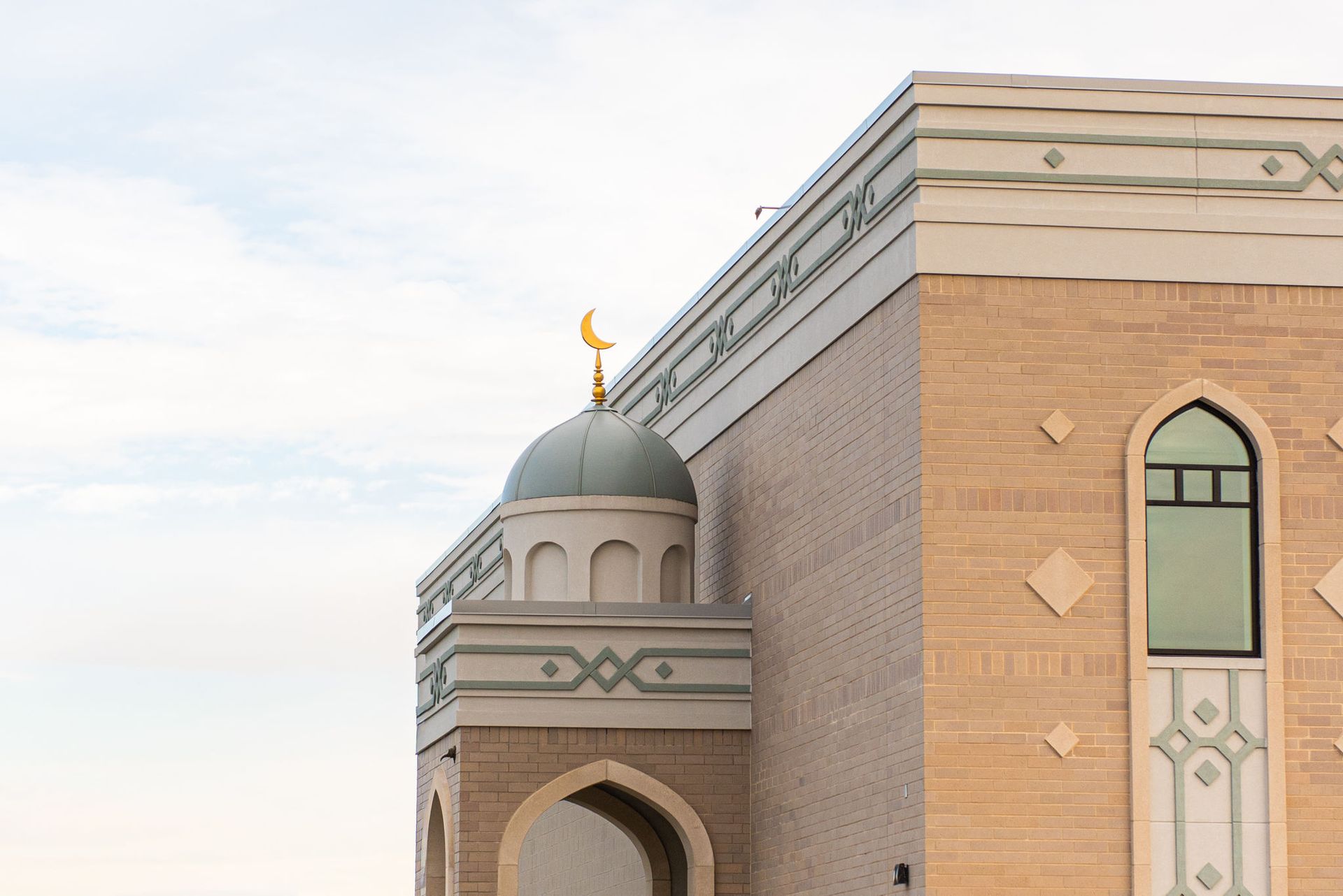 Side view of a mosque with a turquoise dome topped by a crescent moon, tan brick exterior, and arched entryway.