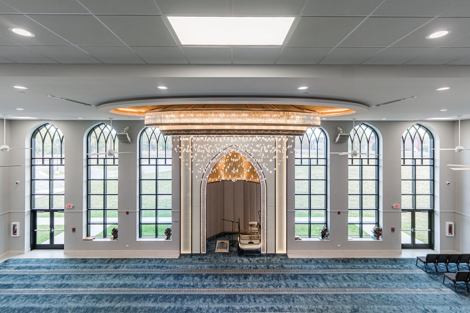 Interior of a mosque with a large blue carpet, tall windows, and ornate chandelier over the mihrab.