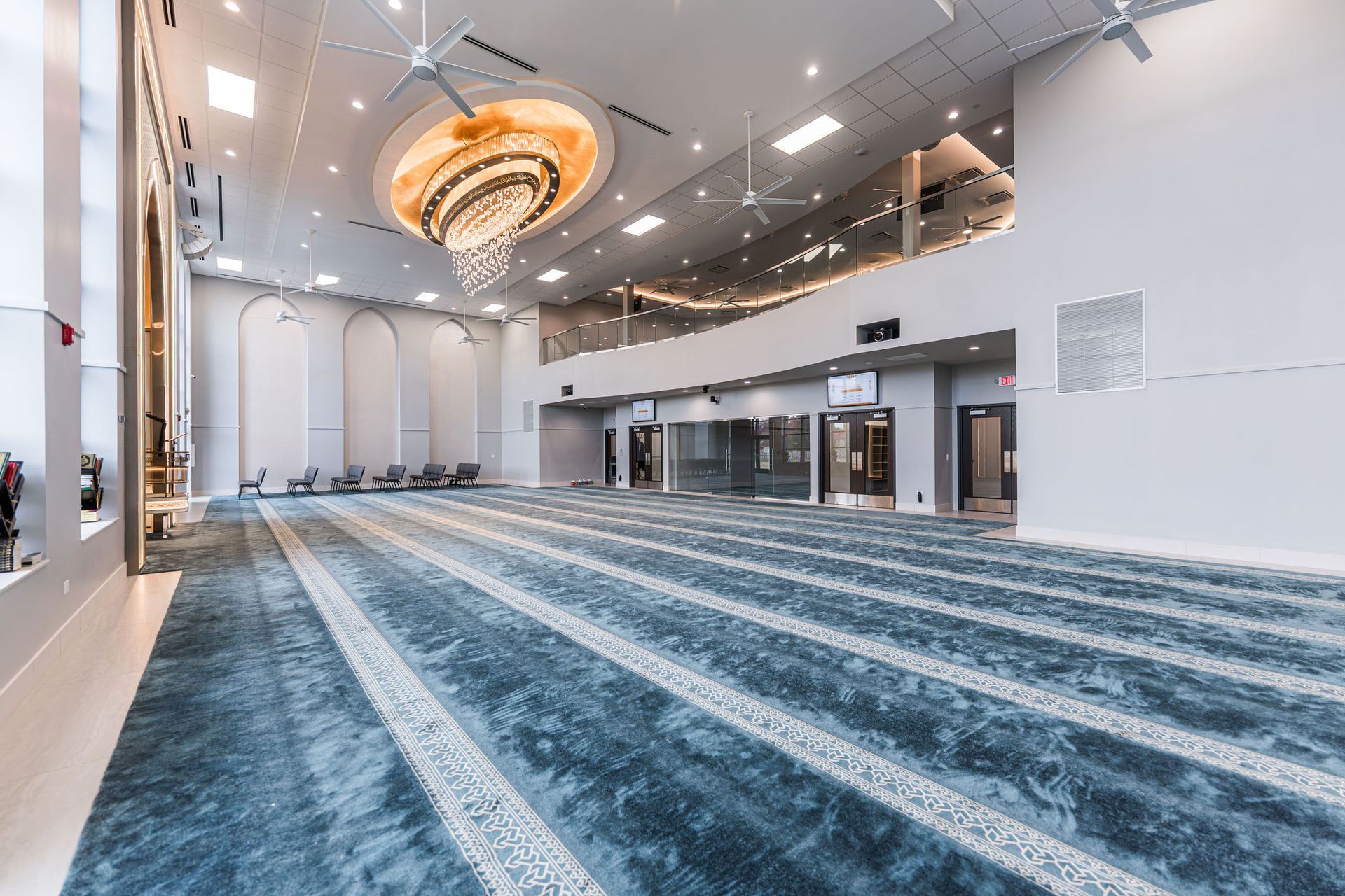 Spacious mosque interior with blue patterned carpet, high ceilings, chandelier, and balcony.