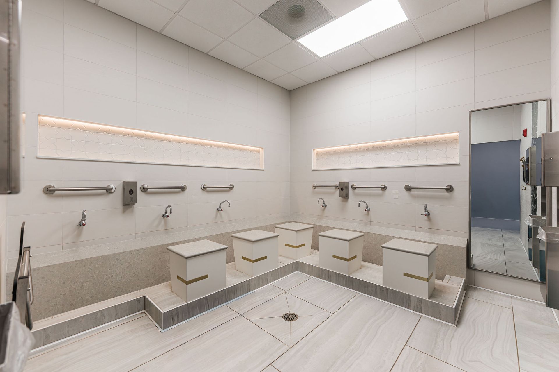 Modern, accessible restroom with multiple sinks, footrests, and a large mirror. Beige and gray color scheme.