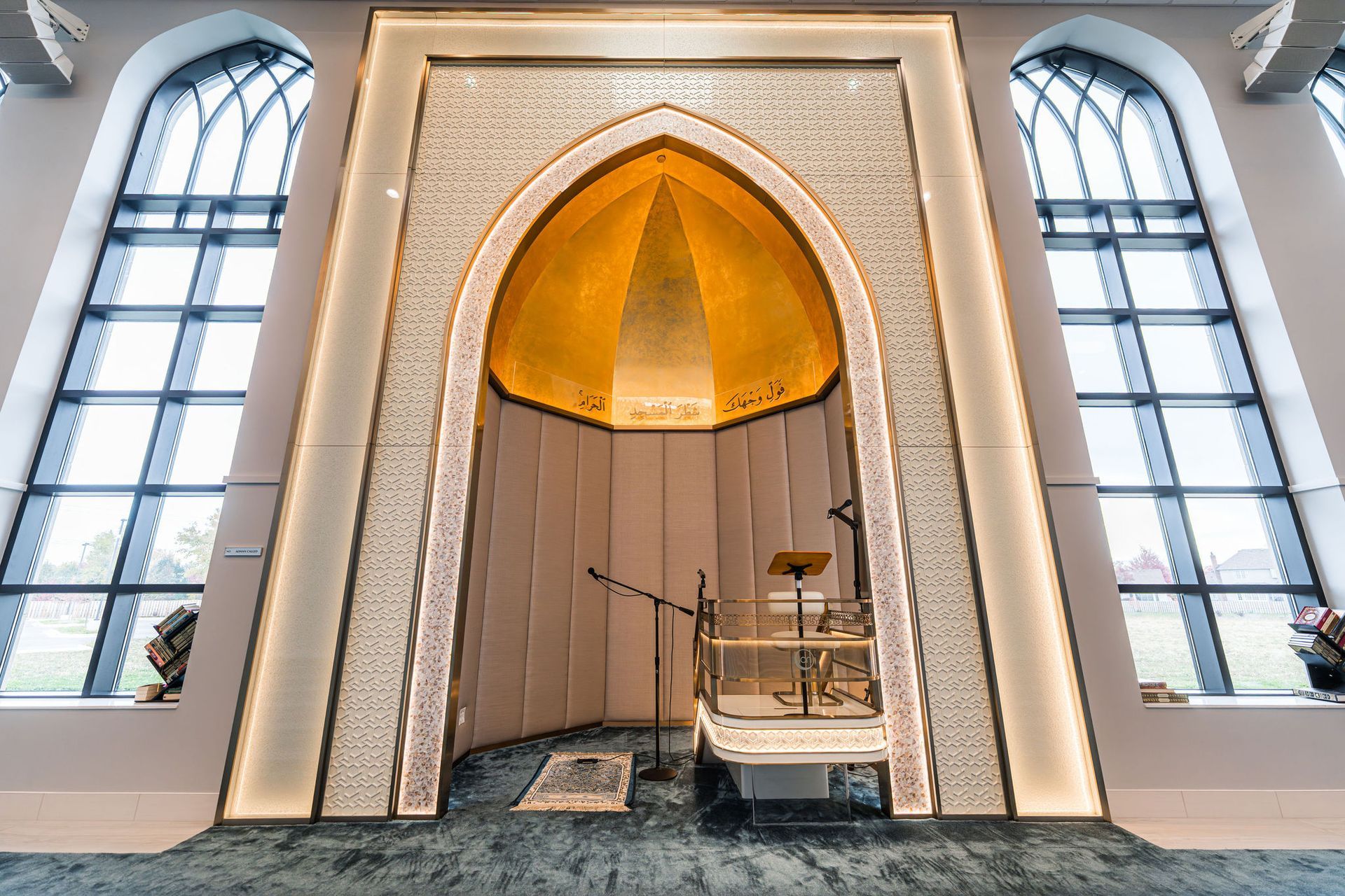 Interior view of a mosque. A gold-domed mihrab is framed by large windows. A lectern stands below.