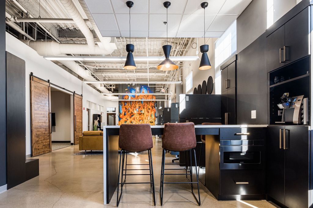 Modern office kitchen with black cabinets, bar stools, and pendant lights.