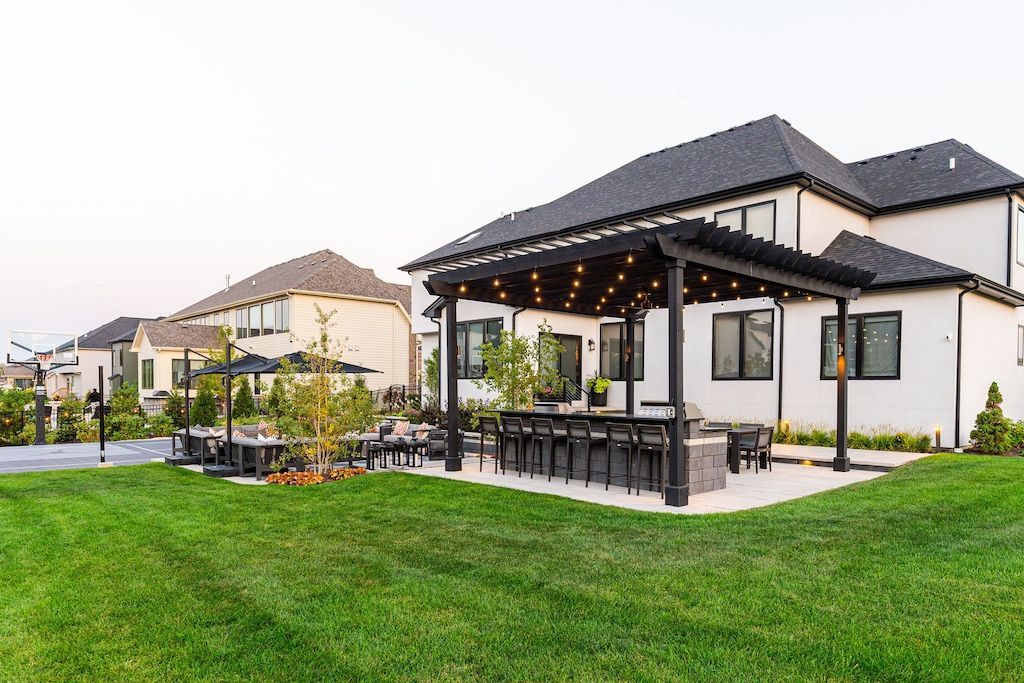 A modern white house with a black pergola and patio, set on a green lawn.