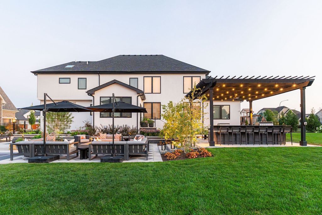 Backyard patio with bar, seating, pergola with string lights, and white house.