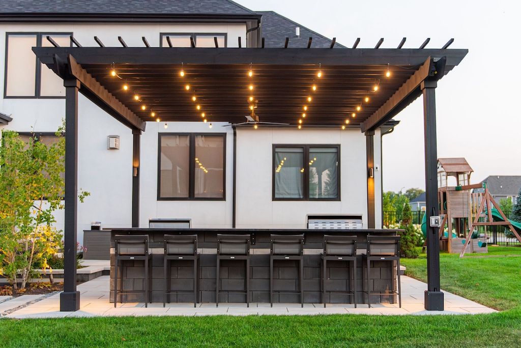 Black pergola with string lights over a bar and stools; white house in the background.