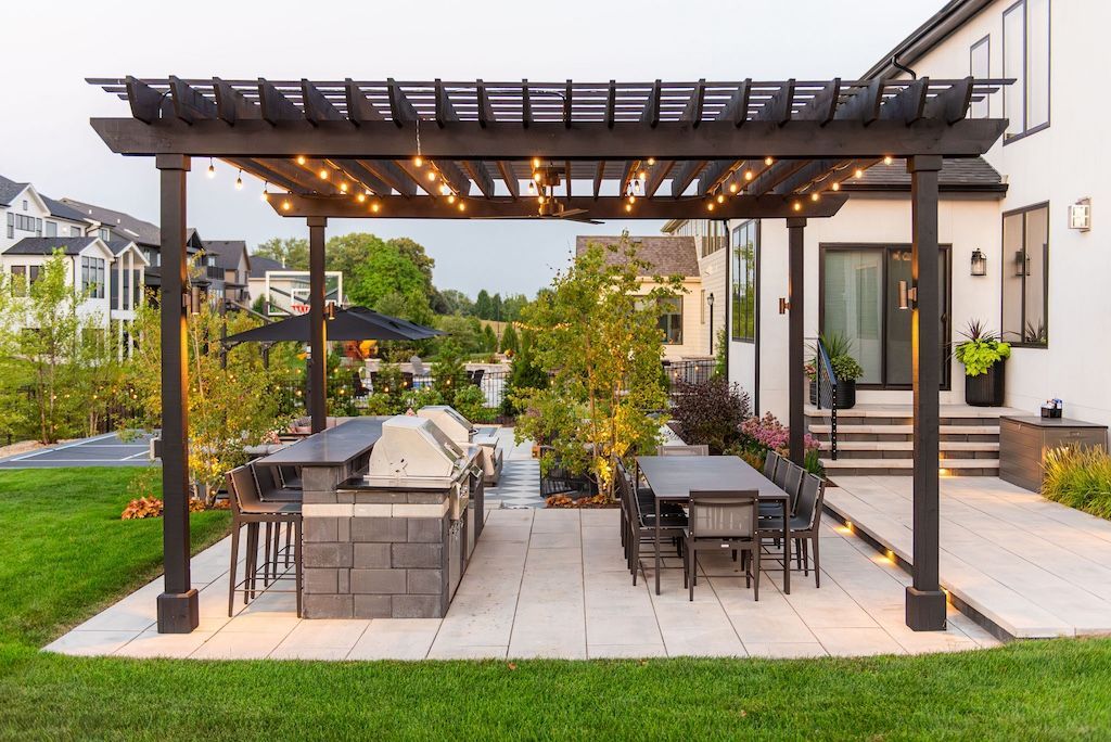 Outdoor kitchen and dining area under a dark pergola with string lights, on a patio next to a house.