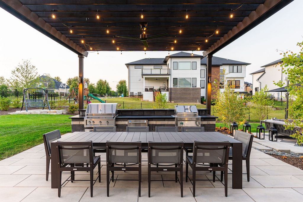 Outdoor dining area with a long table, grill, and patio furniture under a pergola.