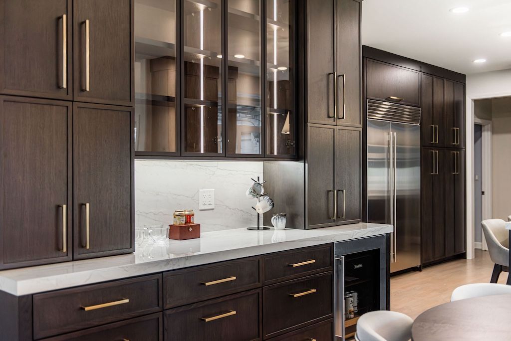 Dark wood cabinetry in a kitchen setting; countertop bar with glassware, wine fridge, and stainless steel refrigerator.