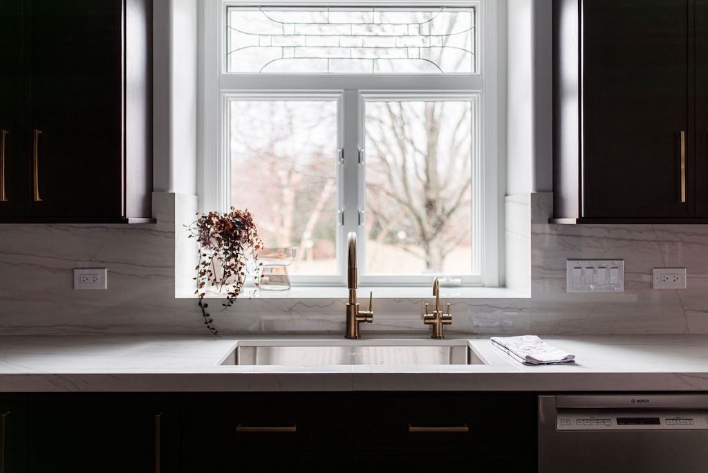 Kitchen sink with a gold faucet, window, and dark cabinets.