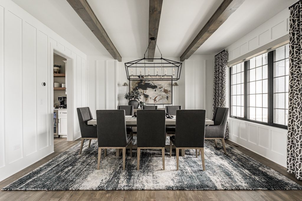 Dining room with a long table and chairs on a patterned rug; beams on the ceiling, black and white window curtains.