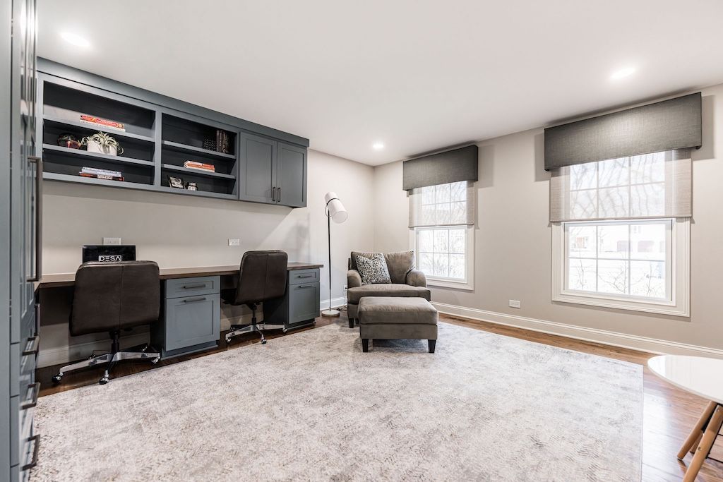 Home office with built-in desk, gray cabinetry, two workstations, armchair, ottoman, and white, fluffy rug.