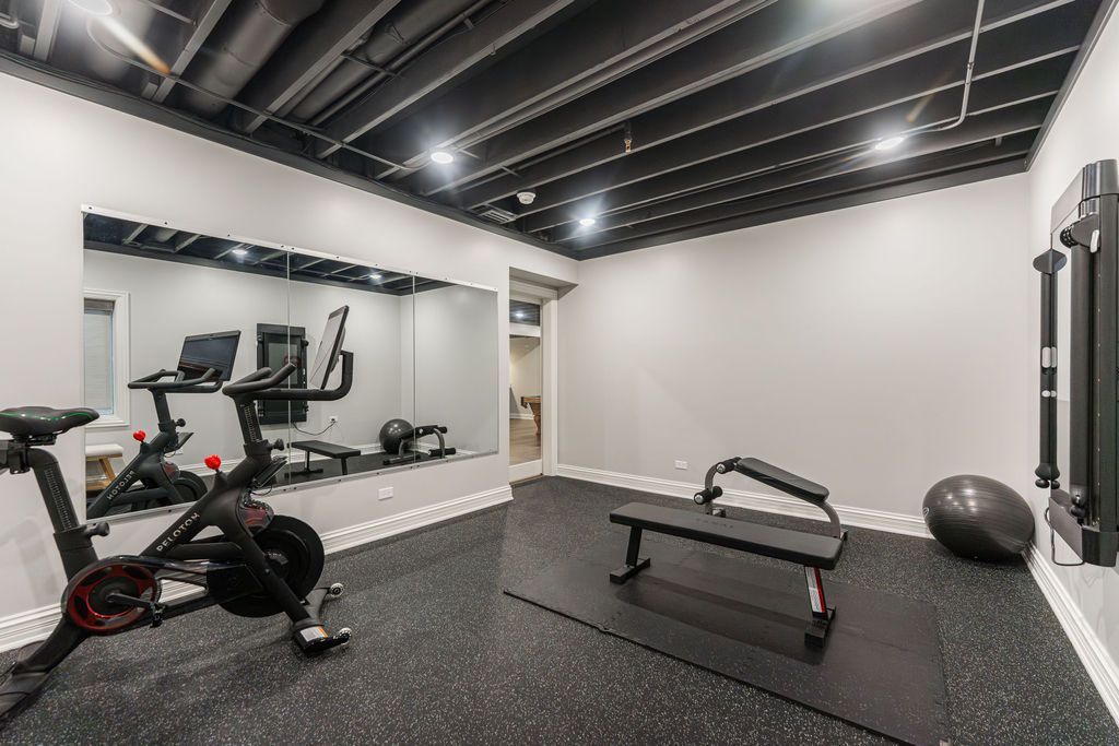 A home gym with a stationary bike, weight bench, exercise ball, mirrored wall, and open-joist black ceiling.