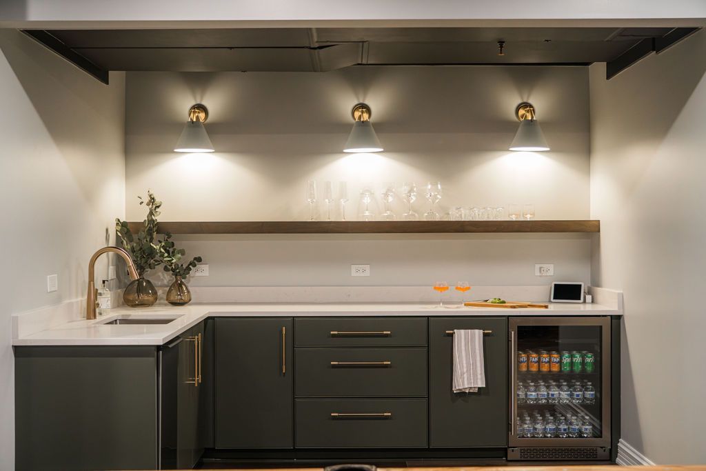 A modern wet bar featuring dark gray cabinets, white countertops, three wall sconces, a floating shelf, and a beverage fridge.
