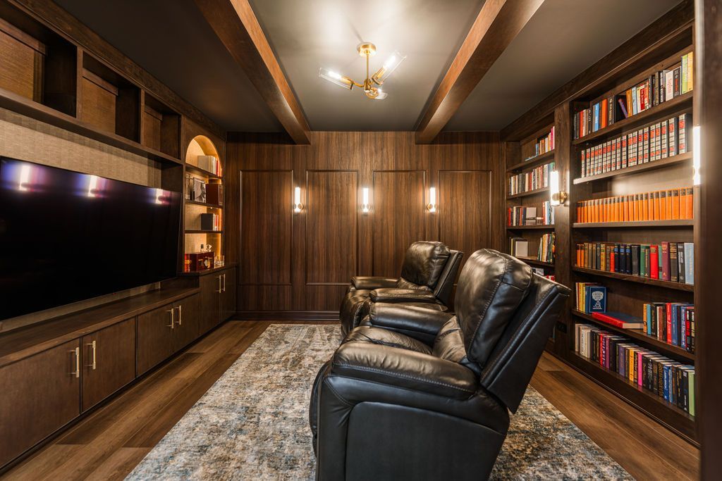 A dark wood home theater with a television, two leather recliners on a patterned rug, and floor-to-ceiling bookshelves.