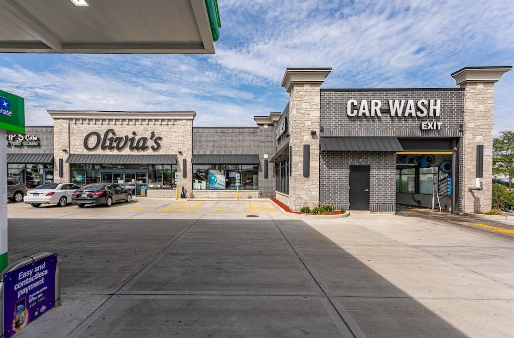Gas station with Olivia's restaurant and car wash. Gray buildings, cars parked in front. Blue sky with clouds.