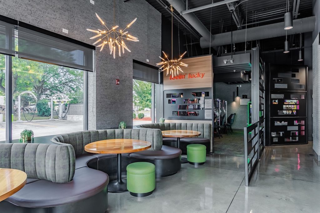 Modern cafe interior with rounded booths, wooden tables, and green stools. Large windows, decorative starburst lights, and polished concrete floor.