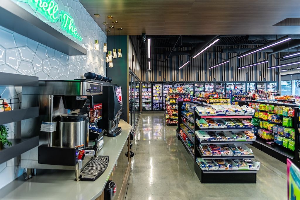 Interior of a convenience store with shelves of products, a coffee station, and a cooler in the background.