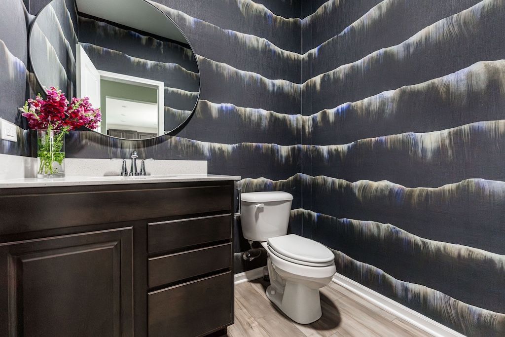 Powder room with dark vanity, patterned wallpaper, round mirror, white toilet, and flowers.