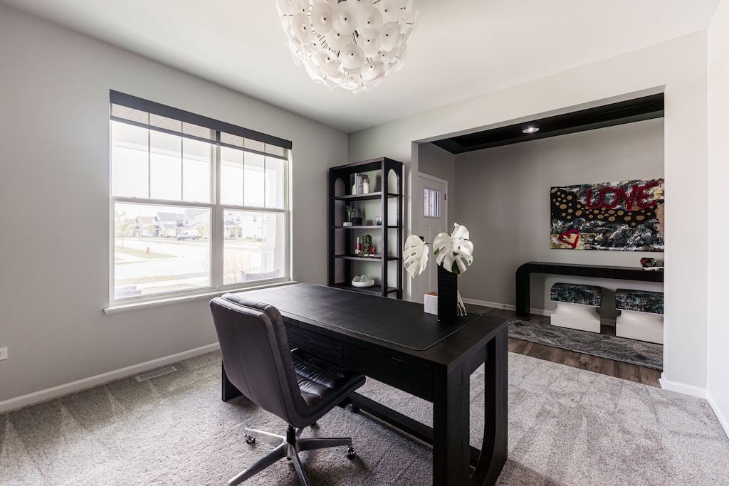 Office with black desk, chair, and shelving against light gray walls and carpet; window on left.
