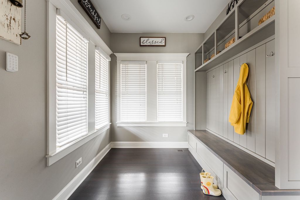 Narrow entryway with gray walls, dark wood floor, built-in bench, yellow jacket, and windows with blinds.