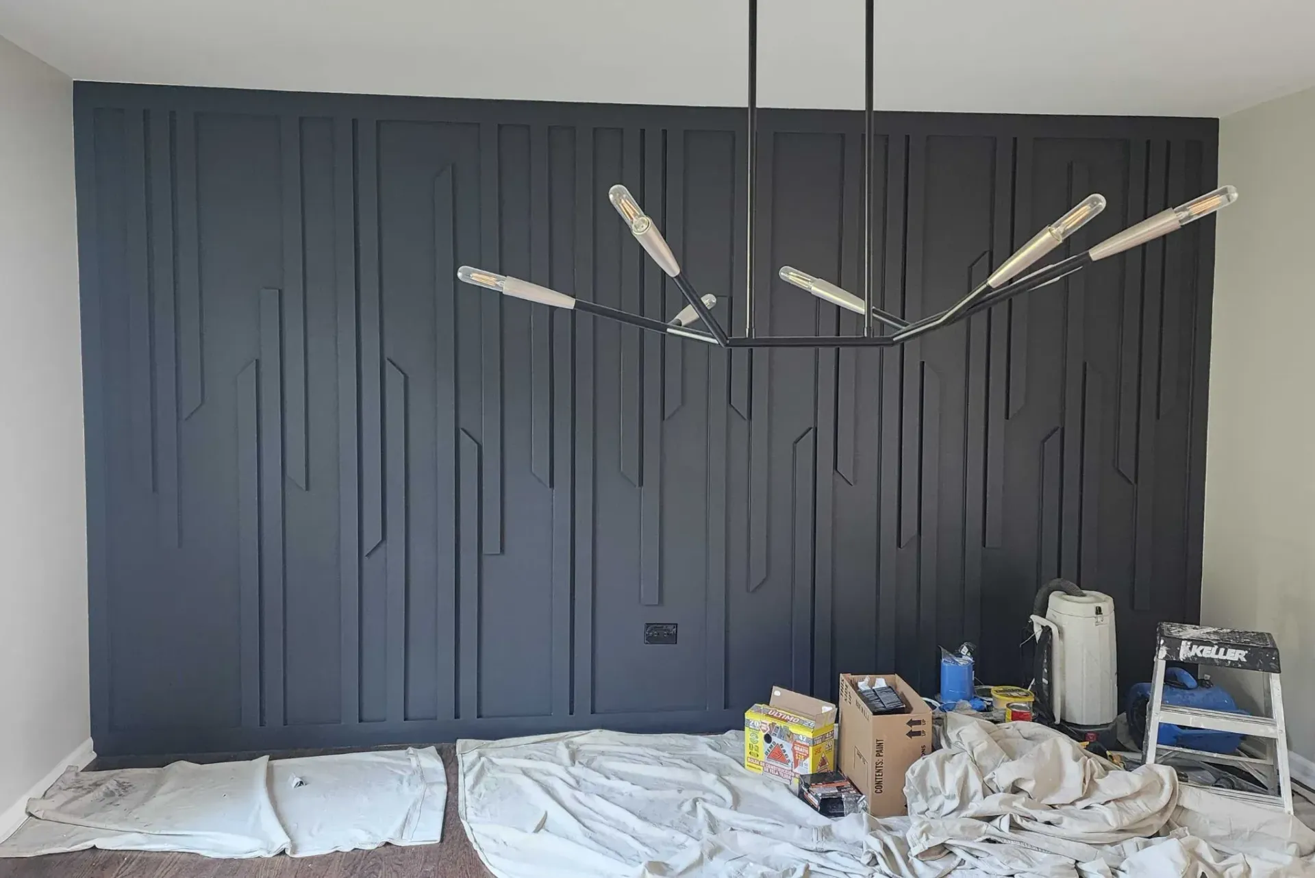 Black accent wall with vertical geometric wood paneling. Modern chandelier. Construction supplies on the floor.