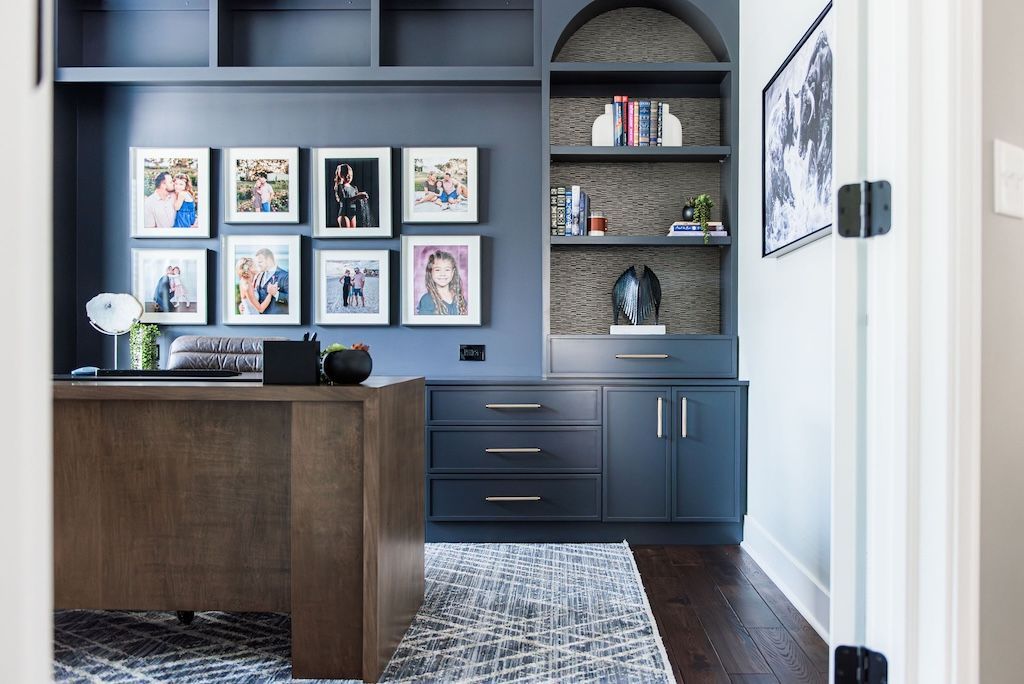Home office with dark blue built-ins, desk, framed photos, and a patterned rug on a hardwood floor.
