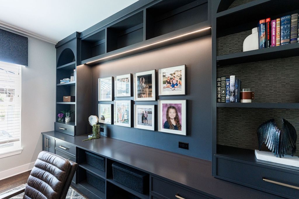 Dark blue home office with built-in desk, shelves, framed photos, and a patterned rug.