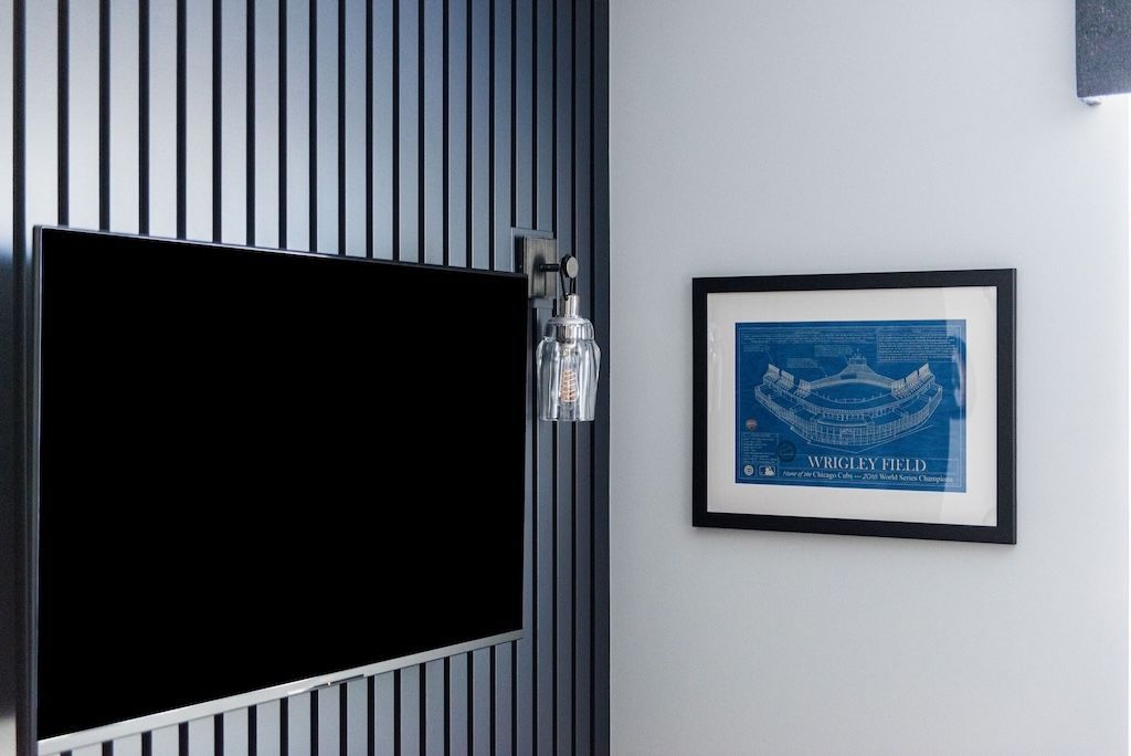 Flat-screen TV on a dark panel wall, with hanging glasses and framed blue print on adjacent white wall.