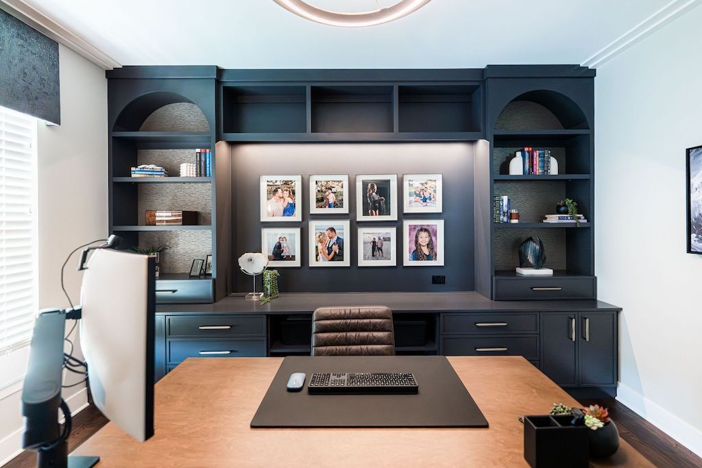 Home office with dark gray built-in shelving, desk, and framed photos. Beige desk and brown leather chair.