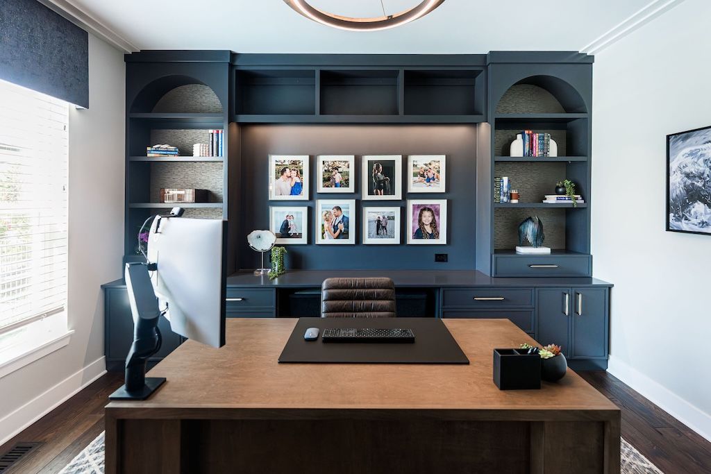 Home office with dark blue built-in desk, shelves, and framed photos. Wooden desk and floor.