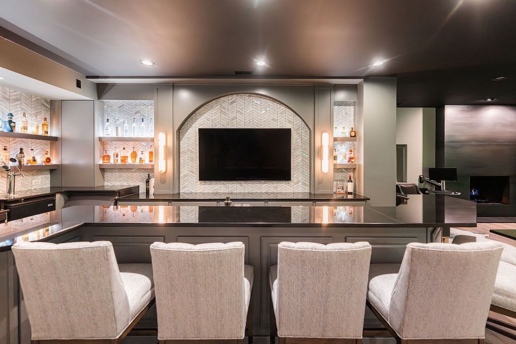 Bar with gray countertop, four light-colored chairs, and shelves of liquor bottles.