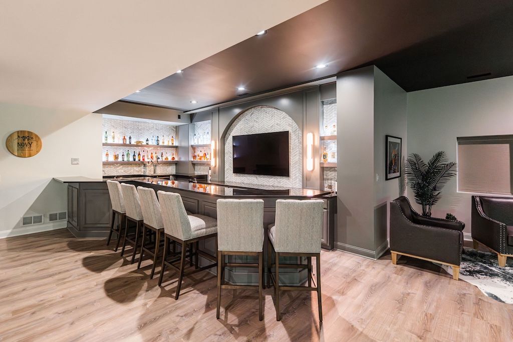 Basement bar with dark ceiling, wood floor, stools, and built-in shelving with bottles.