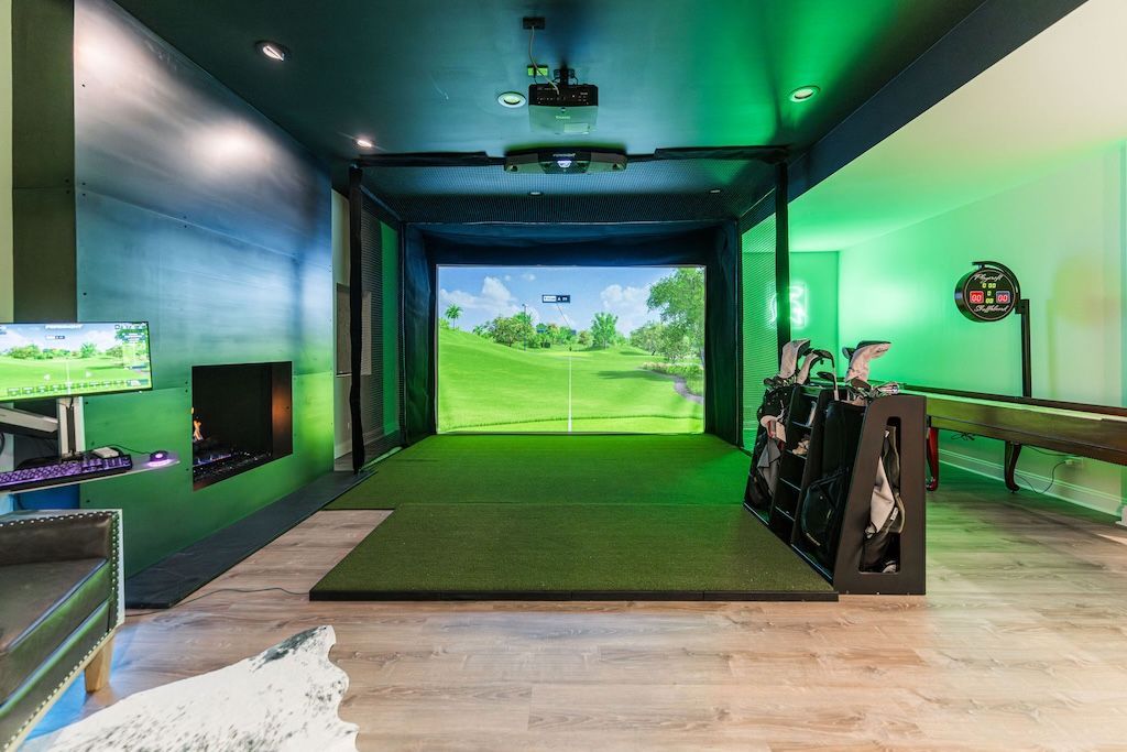 Indoor golf simulator with a green screen, clubs, and putting mat.  Green and blue lighting.