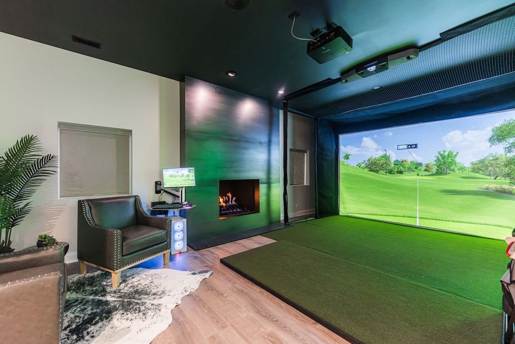Golf simulator room: green screen, turf, fireplace, leather chair, projector, golf course backdrop.