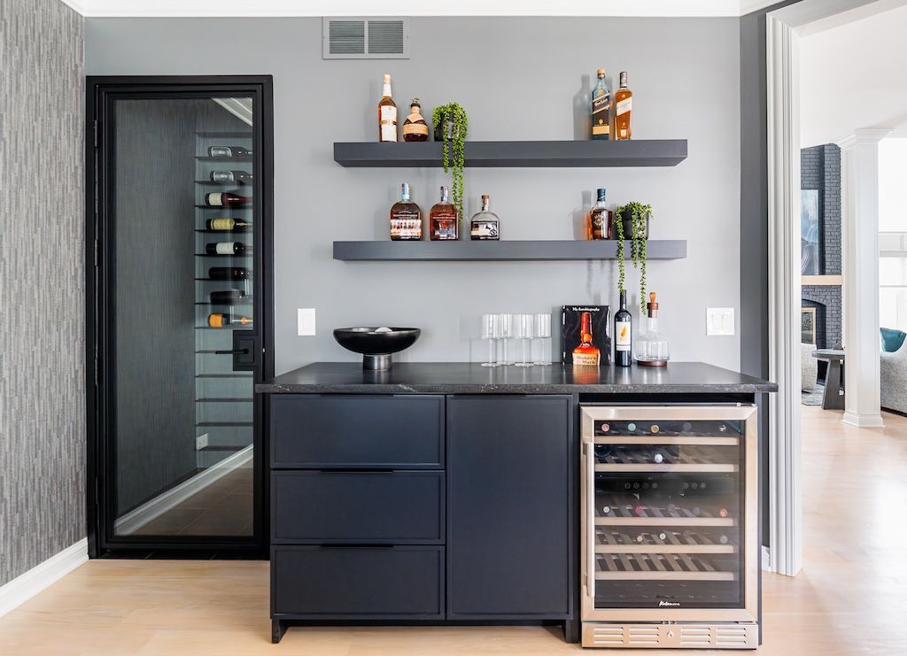 A modern home bar with dark blue cabinetry, wine cooler, floating shelves, and a mirrored wine cellar door.