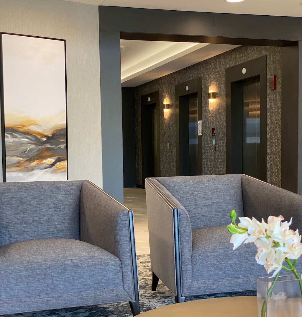 Two gray armchairs in a waiting area; abstract art, elevators in the background.
