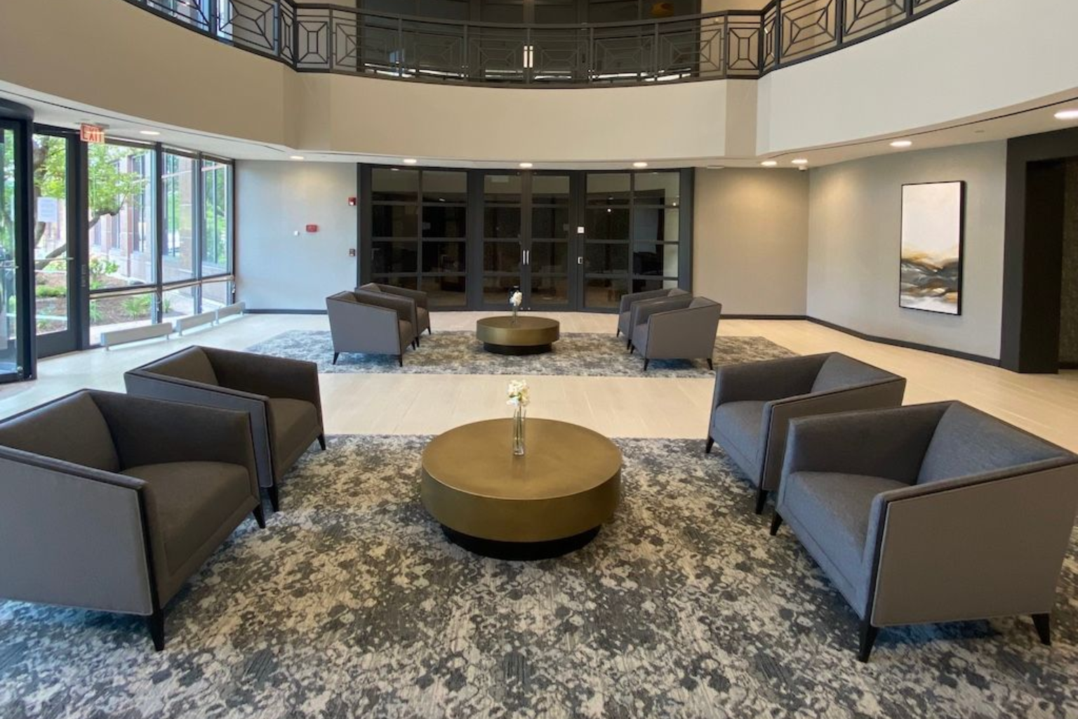 Lobby with gray armchairs around gold ottomans on a patterned rug; large windows and art on walls.