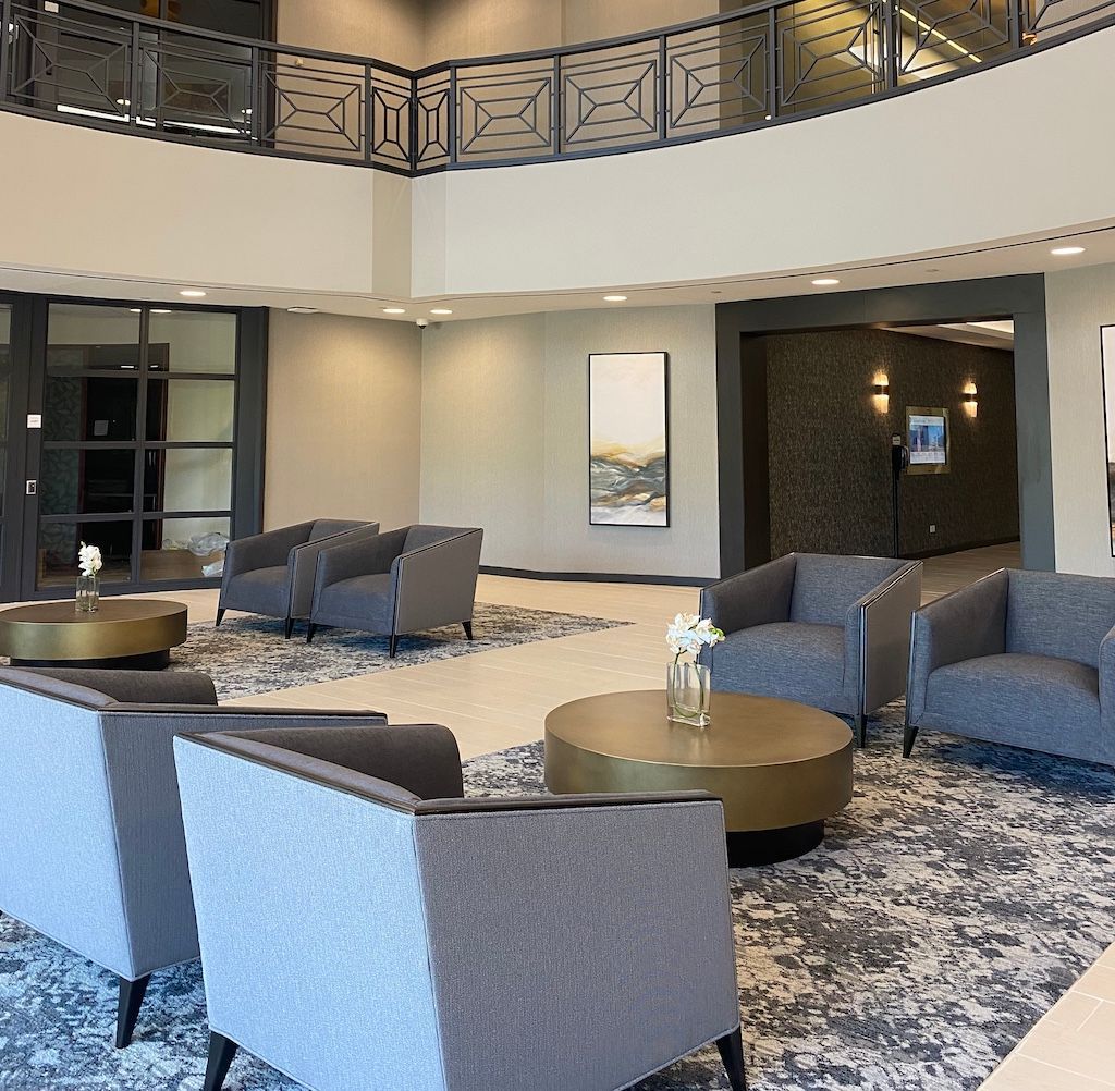 Spacious lobby with gray armchairs, round gold coffee tables, patterned rug, and artwork.