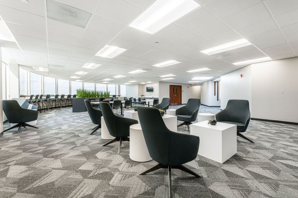 Modern office lounge with gray chairs, white tables, and patterned carpet. Large windows and overhead lighting.