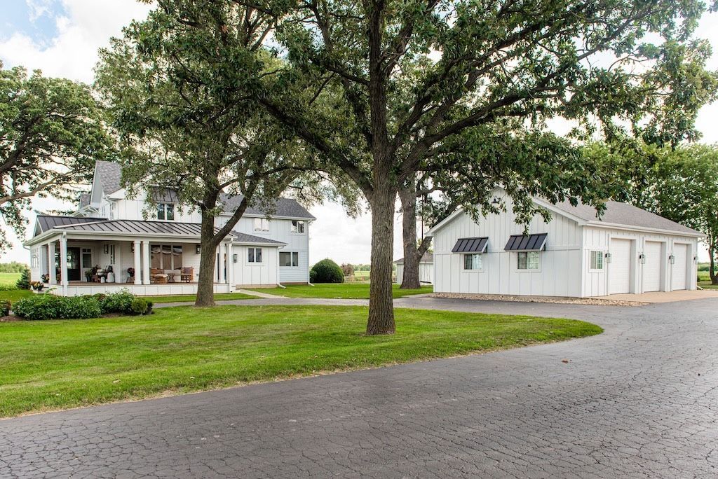 White farmhouse and detached garage on a grassy lot, dark driveway, mature trees.