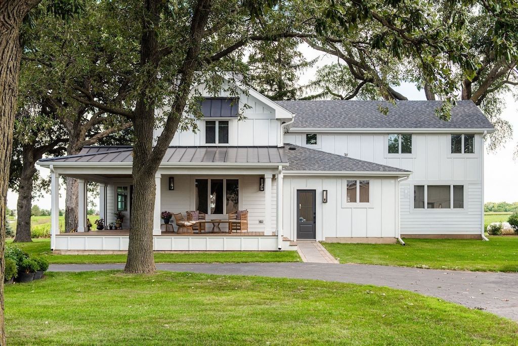 White farmhouse with black accents, porch, and a long driveway on a green lawn.