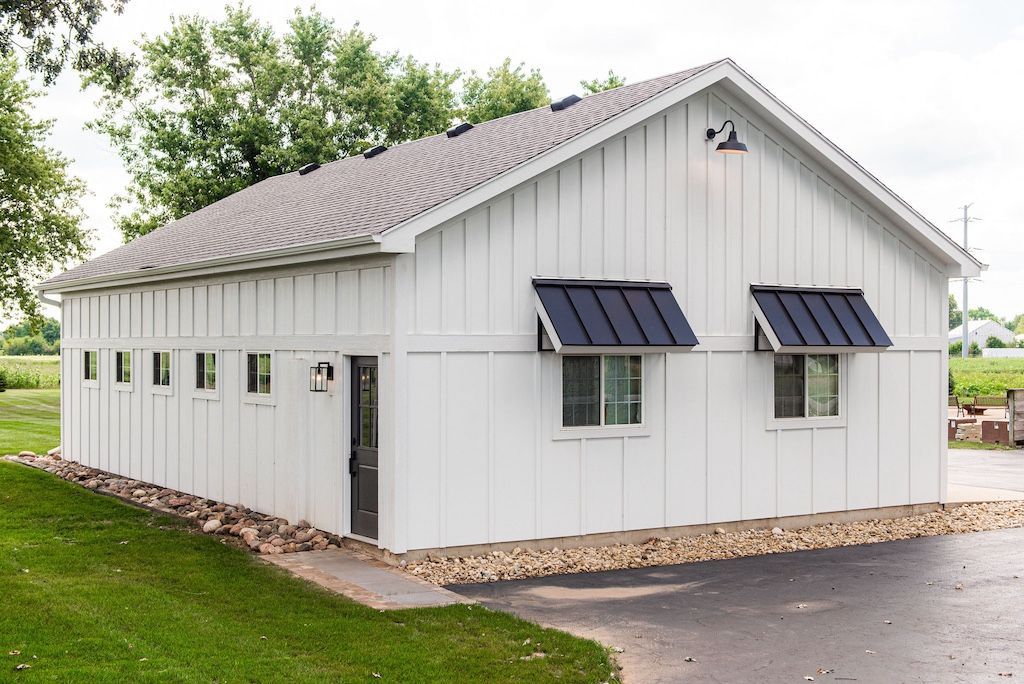 White barn-style building with black awnings and a dark door, on a concrete pad with a gravel base.