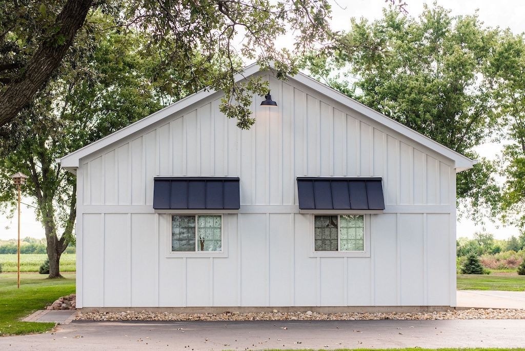 White building with gabled roof, two windows, black awnings, and tree branches overhead.