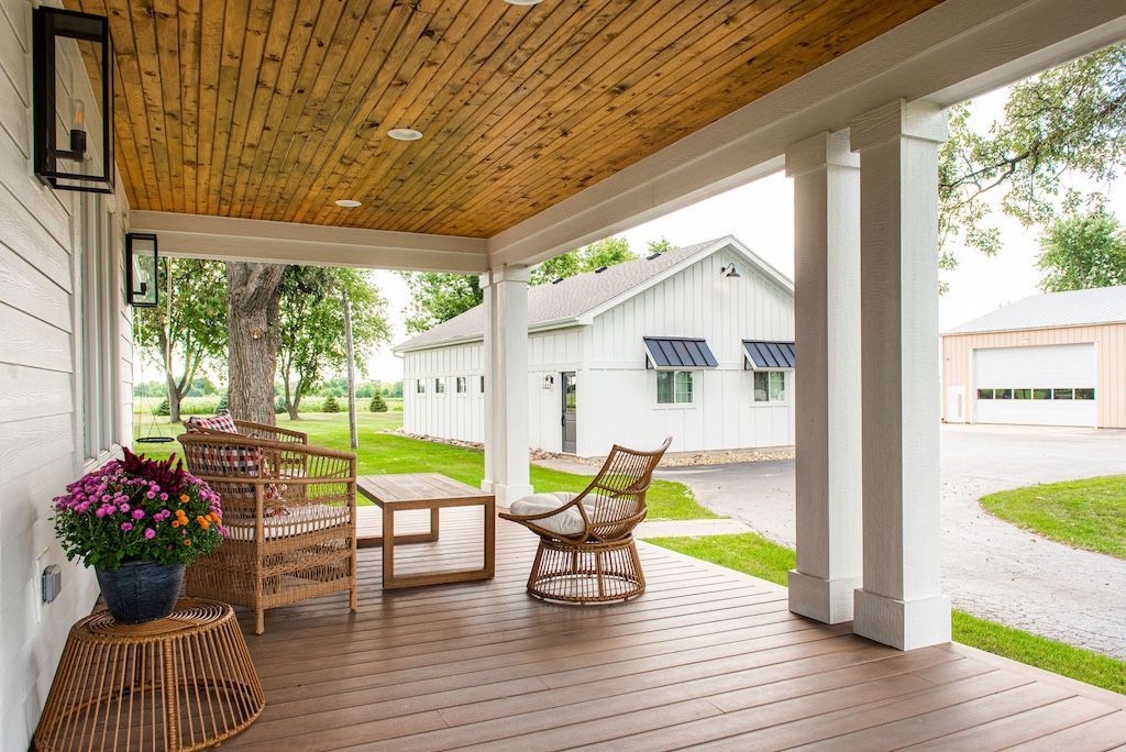 Covered porch with seating; brown ceiling, white columns, lawn, and buildings in the background.