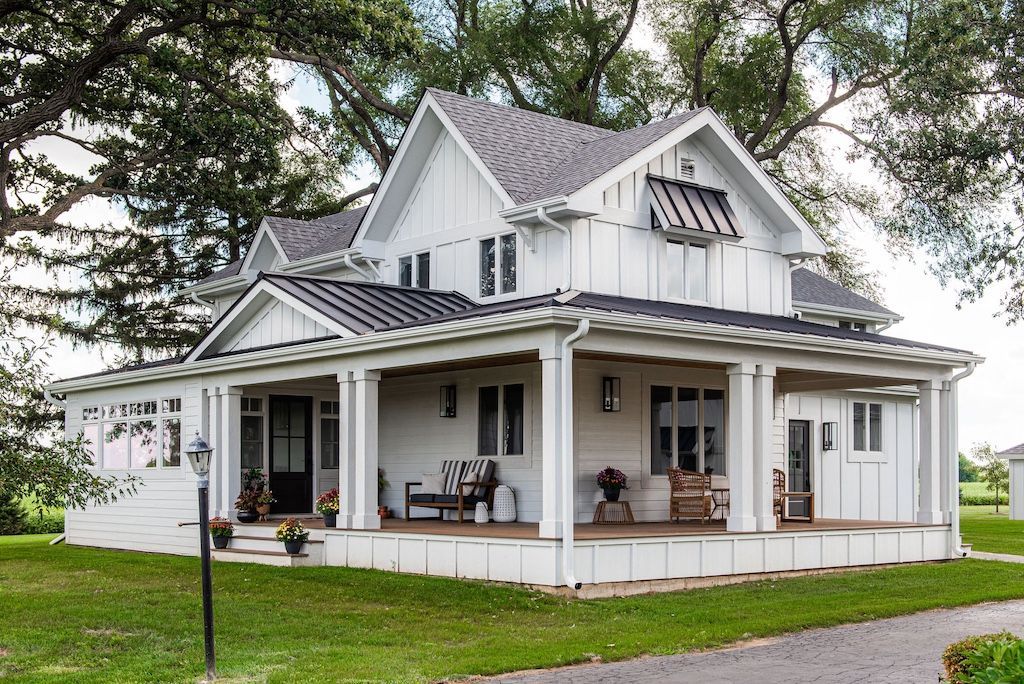 White farmhouse with a wraparound porch and black roof against a green lawn.
