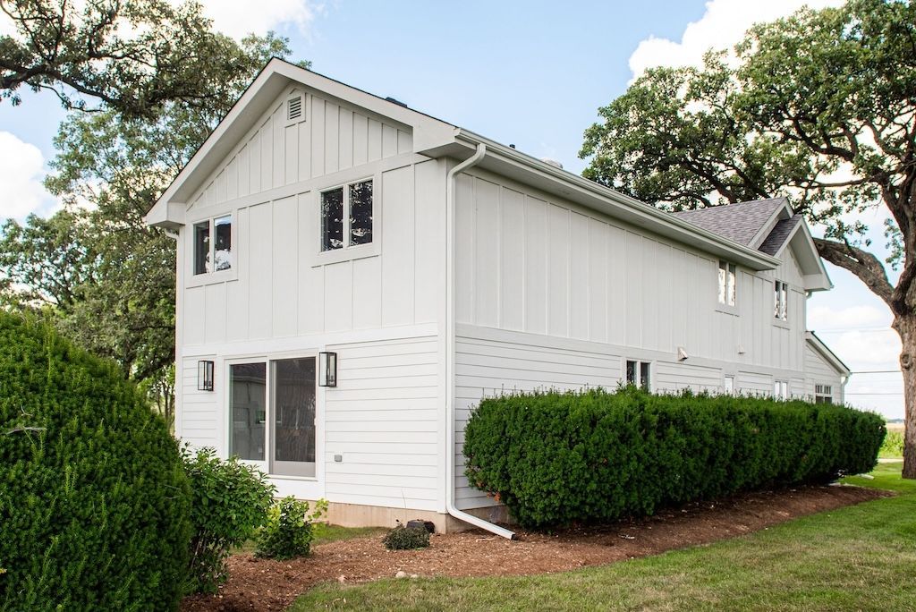 White two-story house with vertical siding and multiple windows, set in a grassy area with trees and shrubs.