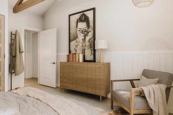 Bedroom with a wooden dresser, a large framed photo, and a gray armchair. The walls are white, and the floor is light-colored wood.
