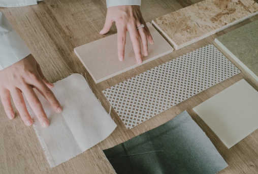 Hands arranging samples of textures and materials on a wooden surface, including fabric and tile.
